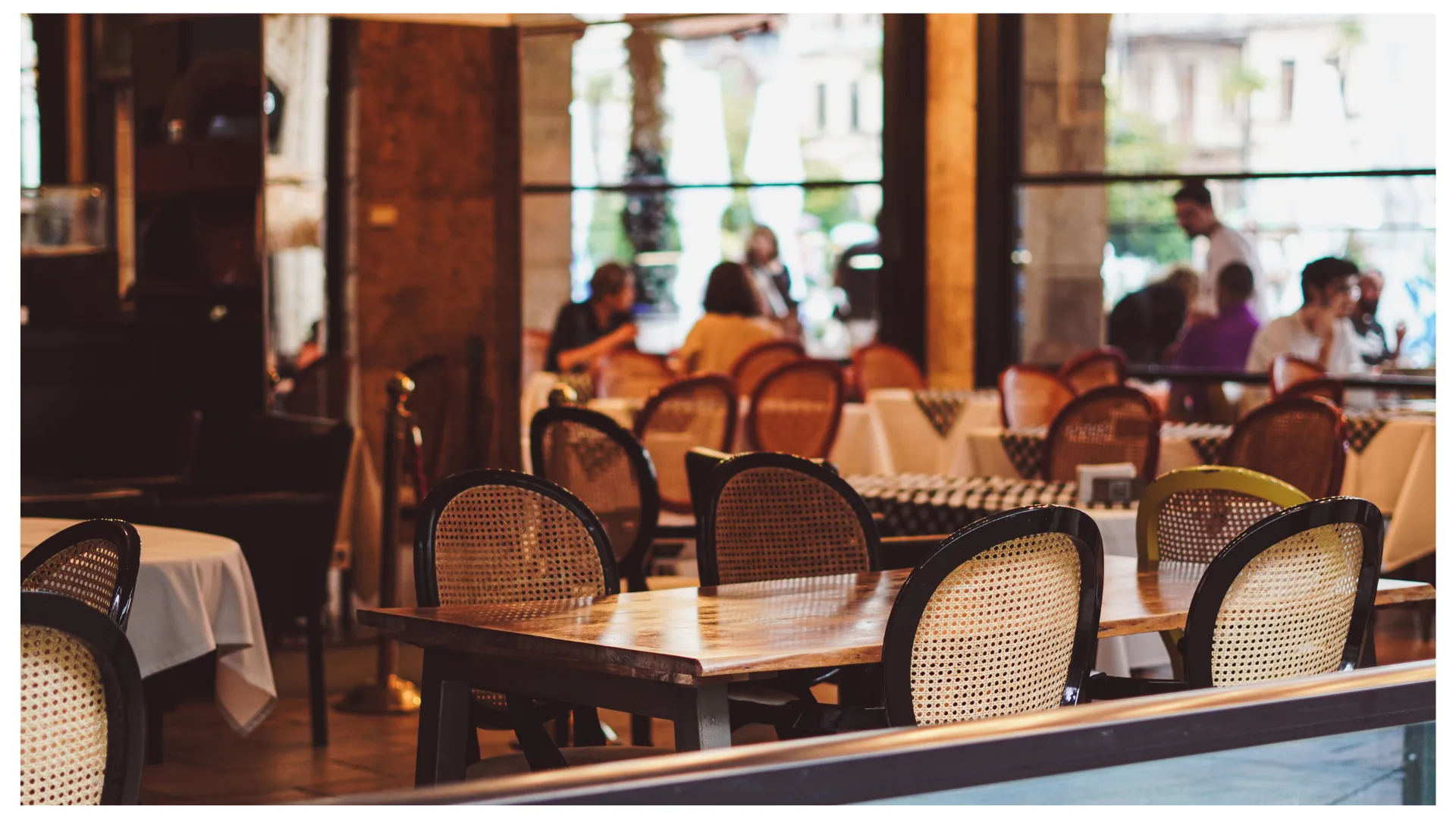 Une salle de restaurant aux chaises cannées, tables dressées et clients attablés en terrasse à l'arrière-plan.